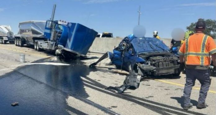 Highway crash scene with a crushed blue car and a blue semi-trailer, fluids spilled on the road, and first responders in orange vests nearby.