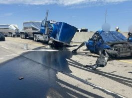 Wrecked blue semi-truck and a severely damaged car on a highway, with a dark fluid spill and responders nearby under a clear sky.