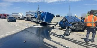 Wrecked blue semi-truck and a severely damaged car on a highway, with a dark fluid spill and responders nearby under a clear sky.