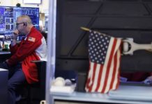 United States strikes Iran for 10th night in a row Man in a red jacket with a US flag patch, seated at a desk with multiple monitors displaying graphs in a control room.