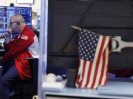 Man in a red jacket with a US flag patch, seated at a desk with multiple monitors displaying graphs in a control room.