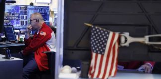 Man in a red jacket with a US flag patch, seated at a desk with multiple monitors displaying graphs in a control room.