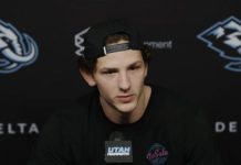 Young man with a backward cap speaks into a microphone at a press conference, backdrop with sponsor logos behind him.
