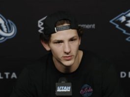 Young man with a backward cap speaks into a microphone at a press conference, backdrop with sponsor logos behind him.