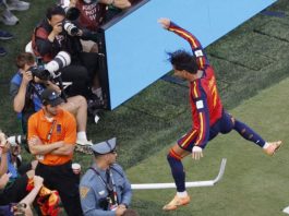 Soccer player in Barcelona kit leaps over sideline barrier as photographers capture the moment from the crowd.
