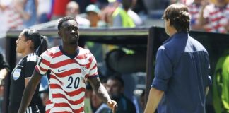 Soccer player wearing a red-and-white striped USA jersey with number 20 walks toward the sideline, with a man in a blue shirt nearby.