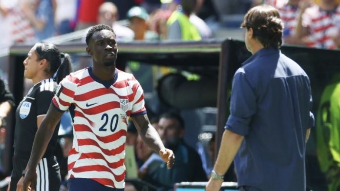 World-Cup-USA-survives-Bosnia-Herzegovina-loses-Balogun-for-next-game 2 Soccer player wearing a red-and-white striped USA jersey with number 20 walks toward the sideline, with a man in a blue shirt nearby.