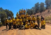 Babylon Fire 95% contained at 107K acres Group of smiling workers in yellow shirts and helmets posing on a dirt trail in a sunny, hilly landscape.