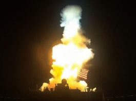 Nighttime rocket launch with a bright orange flame and large plume of smoke rising from a launch pad, with an American flag visible near the base.