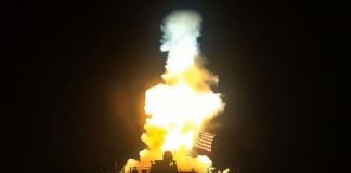 Nighttime rocket launch with a bright orange flame and large plume of smoke rising from a launch pad, with an American flag visible near the base.