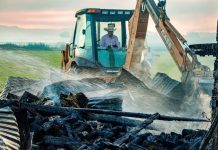 Construction equipment clearing charred debris; a worker in a hat watches from the excavator cab as water sprays over the rubble.