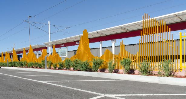 Sunny exterior of a station with tall yellow metal sculptures along the curb and a parking area in the foreground