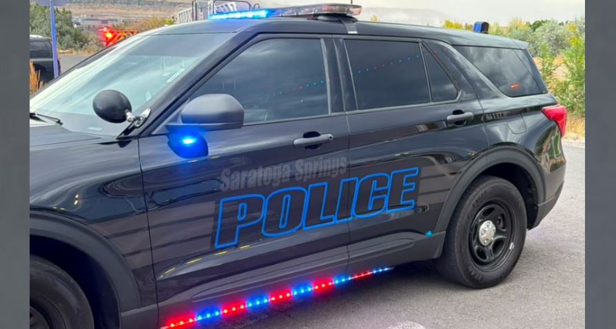 Black police SUV with blue'POLICE' lettering and flashing red/blue lights along the side, parked on a street scene.