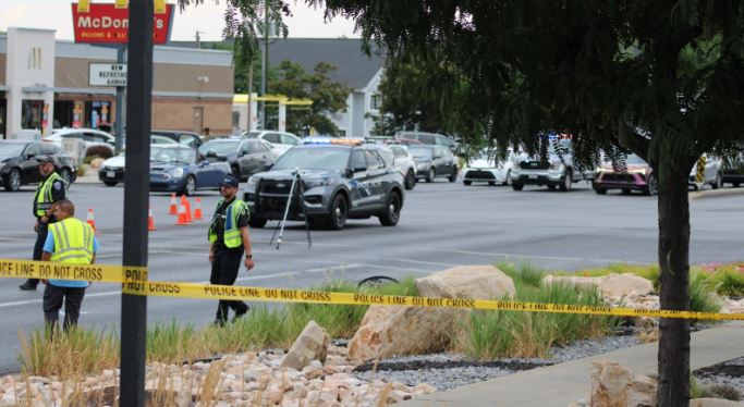 Police officers set up yellow'Police Line Do Not Cross' tape across a parking lot while a patrol car and other vehicles are in the background near a shopping center.