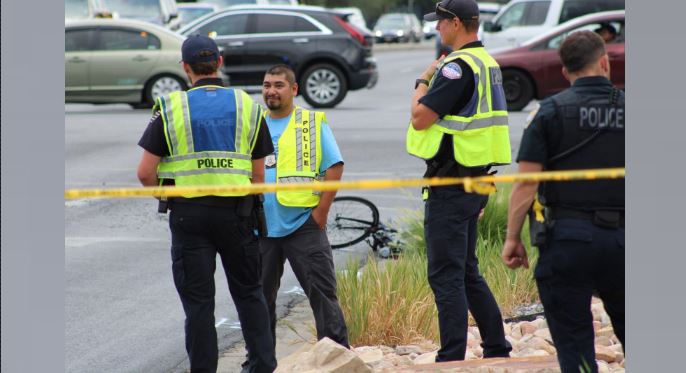 Police officers in high-visibility vests stand behind yellow caution tape at a roadway crime scene; a man in a blue shirt stands nearby with a bicycle on the ground and cars in the background.