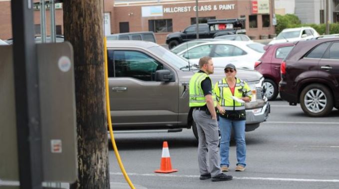 Two people in high-visibility vests talking beside a gray pickup in a parking lot; an orange cone sits nearby.