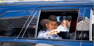 Elderly man wearing a brown cowboy hat and glasses waves from the window of a blue car.
