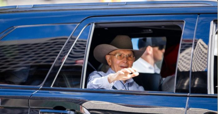 Elderly man wearing a brown cowboy hat and glasses waves from the window of a blue car.