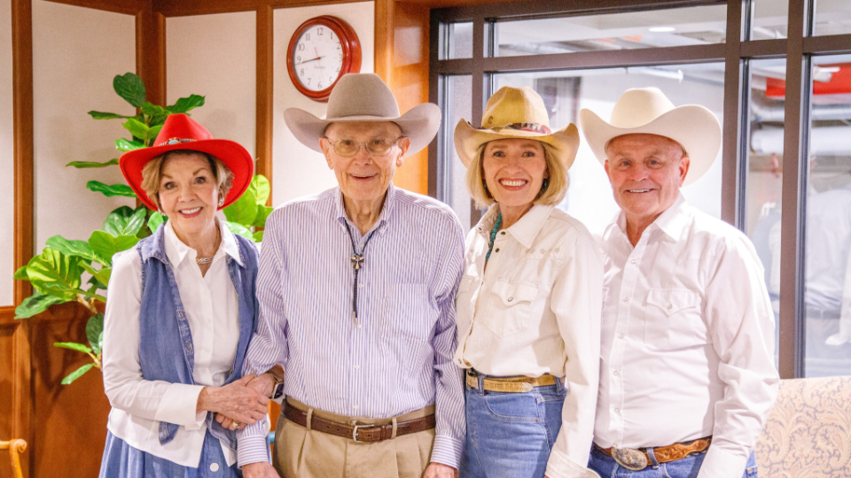 Four seniors in cowboy hats stand together in an office lobby, smiling for the photo.